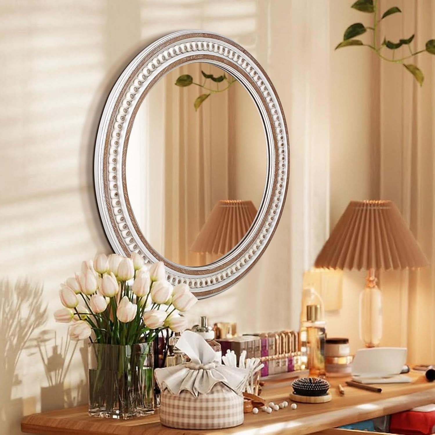 Round decorative mirror on a wall with a table below holding a vase of flowers, books, and a lamp.