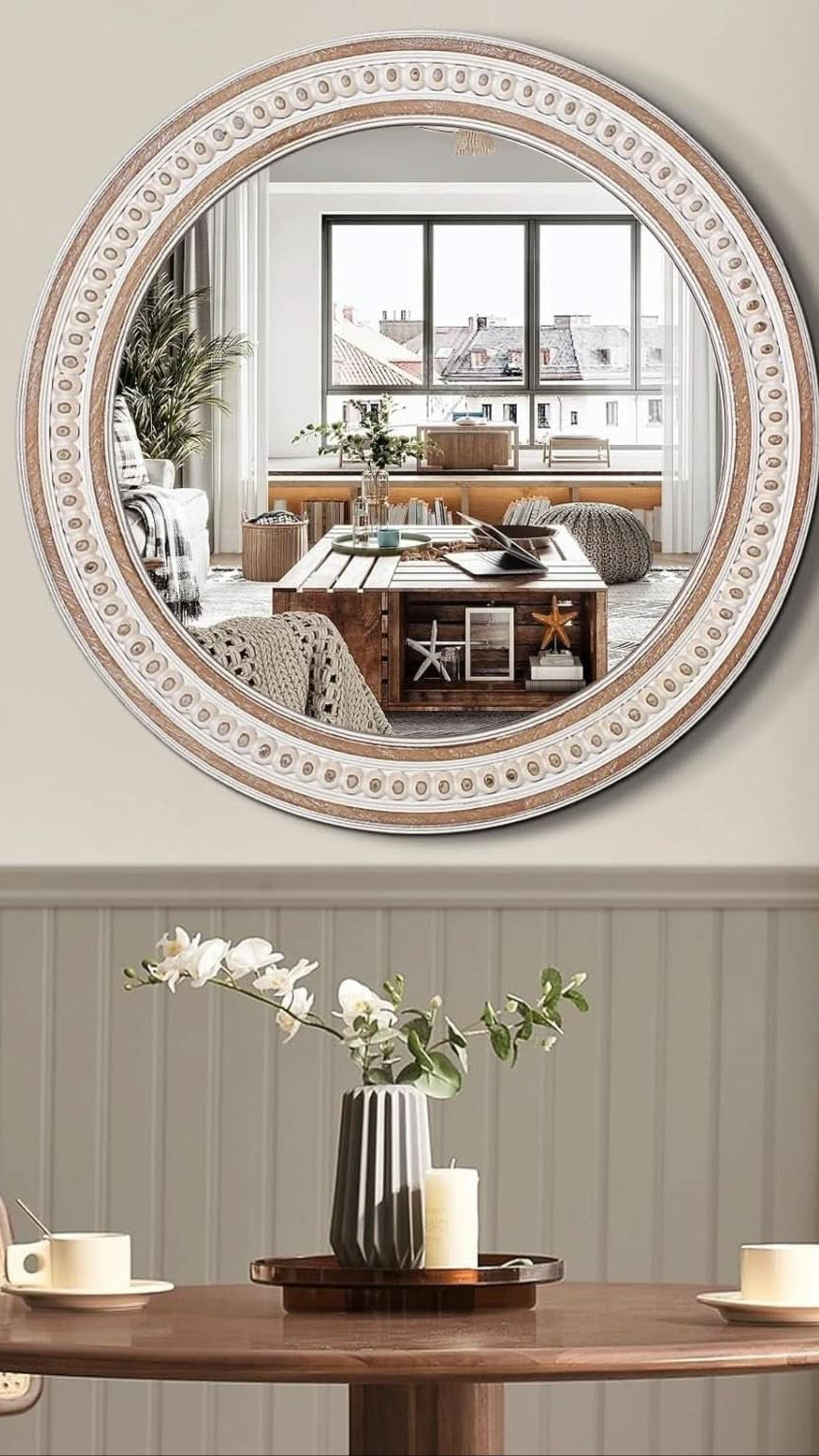 Round decorative mirror with intricate frame on a wall above a table with a vase and candles.