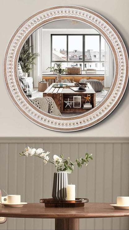 Round decorative mirror with intricate frame on a wall above a table with a vase and candles.