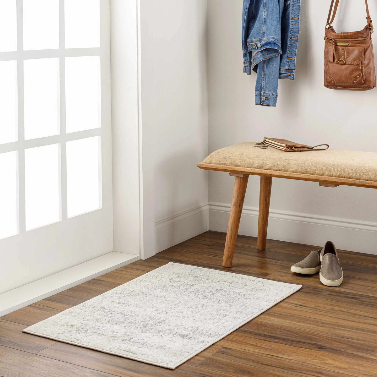 A rectangular light beige entryway rug with a subtle distressed pattern, shown on a dark wood floor in a bright, modern entryway with a wooden bench, shoes, and wall accessories.