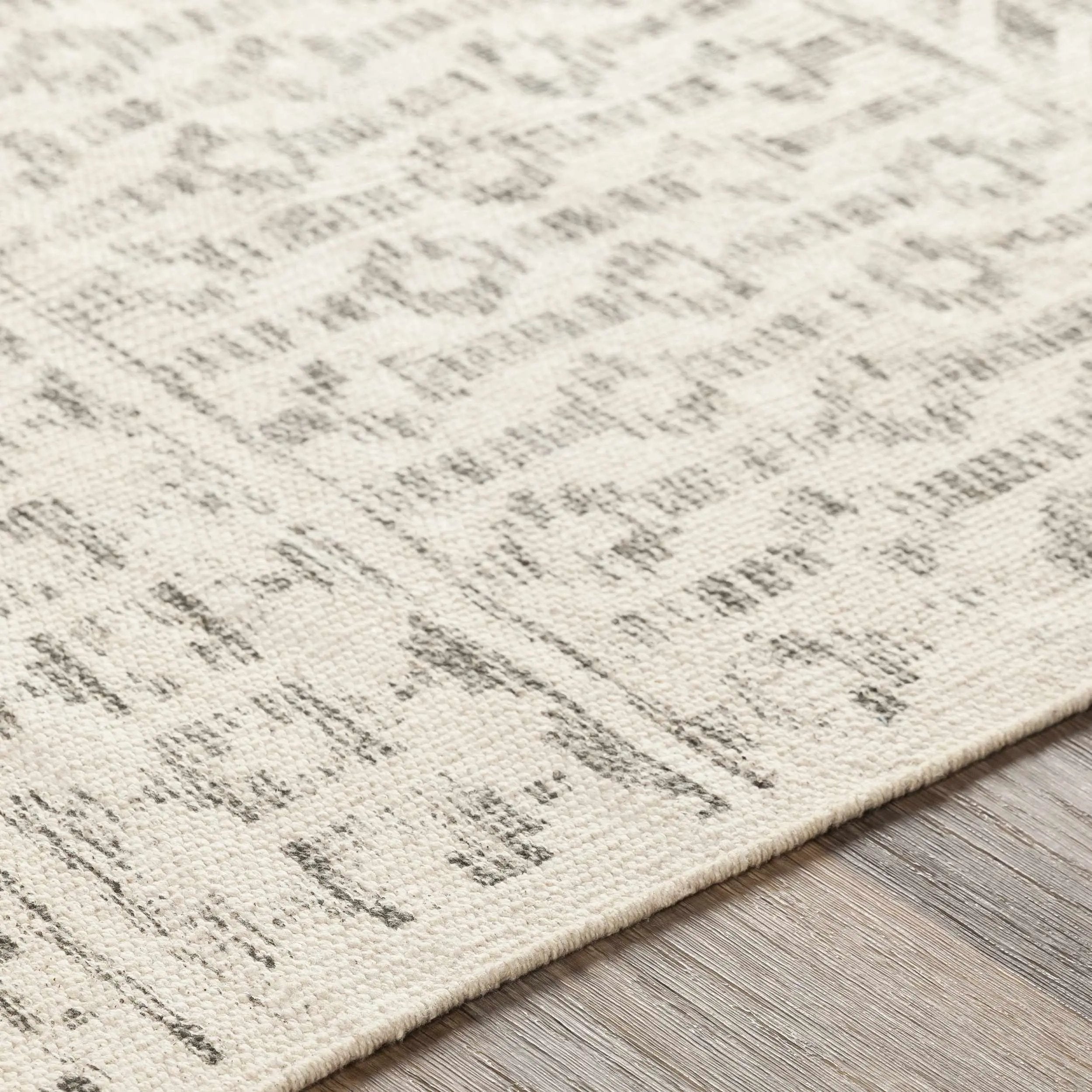 Close-up of a textured area rug with a distressed, abstract pattern in cream and gray, shown on a light wood floor.