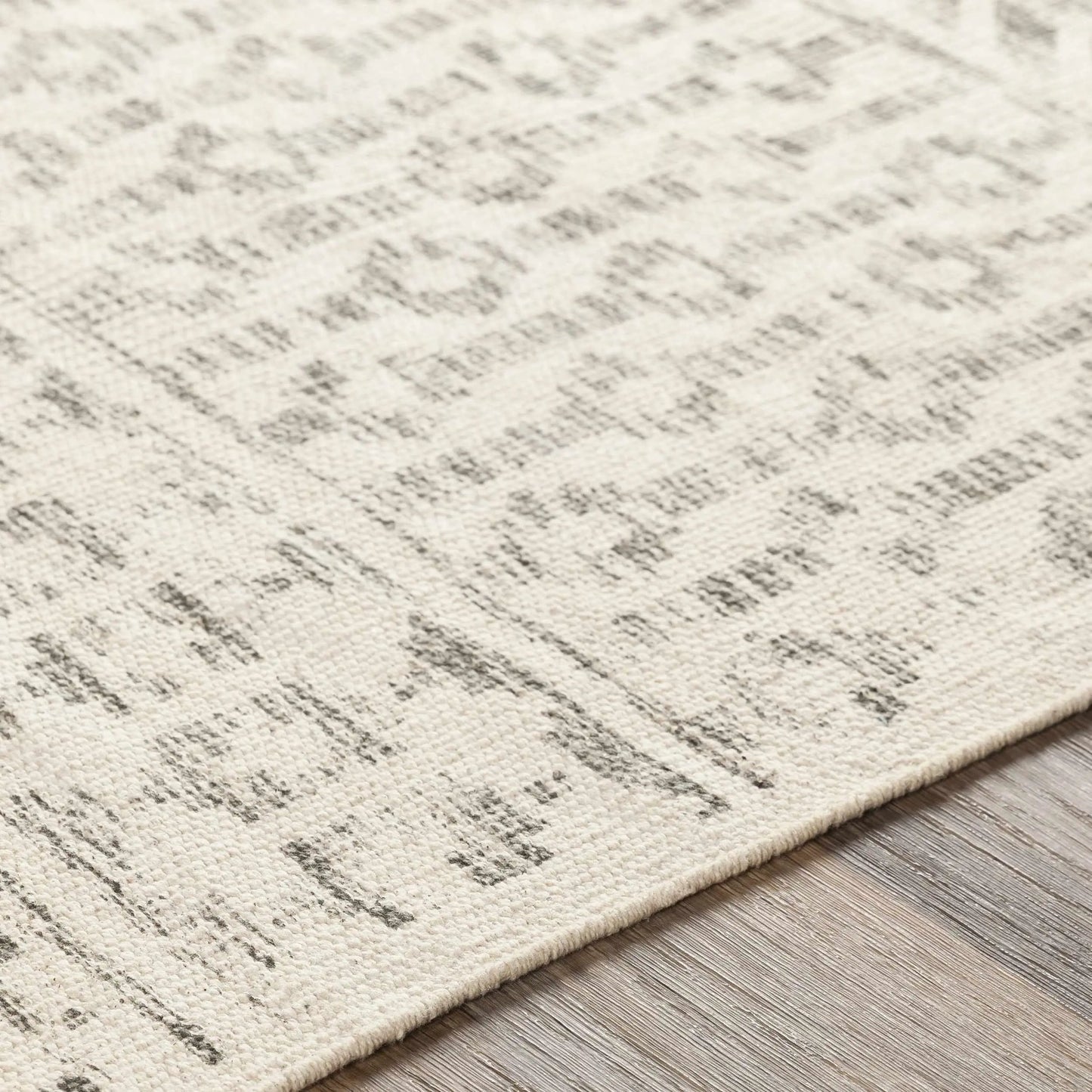 Close-up of a textured area rug with a distressed, abstract pattern in cream and gray, shown on a light wood floor.