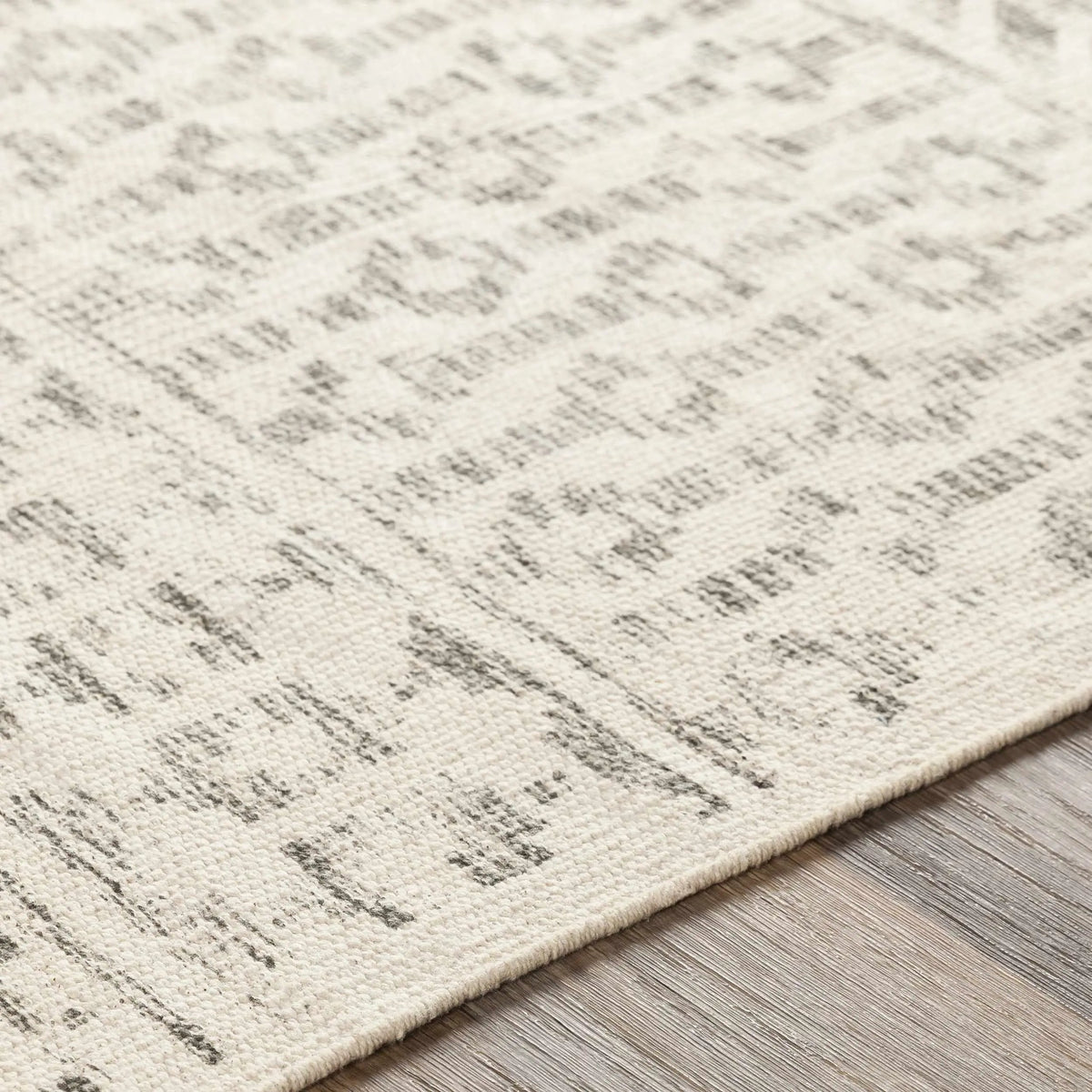 Close-up of a textured area rug with a distressed, abstract pattern in cream and gray, shown on a light wood floor.